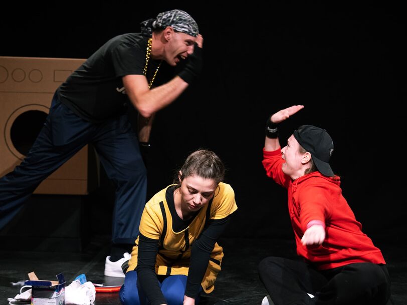 Drei, anhand ihrer Kleidung offensichtlich als Jugendliche zu definierende Personen sitzen bzw. stehen auf einer Bühne, im Hintergrund sind Waschmaschinen zu erkennen. Ein Mädchen malt mit Kreide auf den Boden, die beiden anderen klatschen sich ab (High Five). 