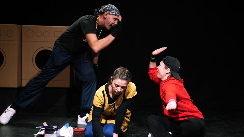 Drei, anhand ihrer Kleidung offensichtlich als Jugendliche zu definierende Personen sitzen bzw. stehen auf einer Bühne, im Hintergrund sind Waschmaschinen zu erkennen. Ein Mädchen malt mit Kreide auf den Boden, die beiden anderen klatschen sich ab (High Five).