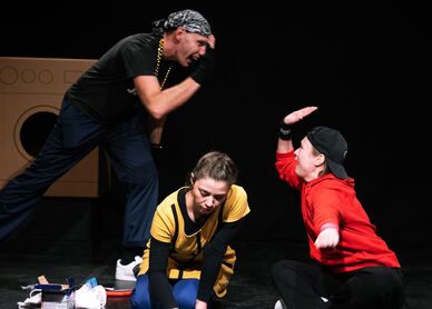 Drei, anhand ihrer Kleidung offensichtlich als Jugendliche zu definierende Personen sitzen bzw. stehen auf einer Bühne, im Hintergrund sind Waschmaschinen zu erkennen. Ein Mädchen malt mit Kreide auf den Boden, die beiden anderen klatschen sich ab (High Five). 