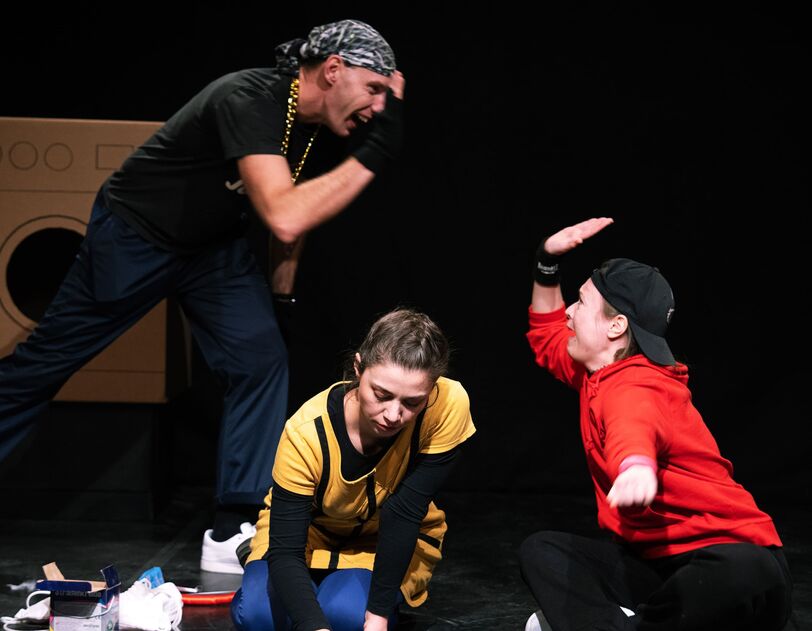 Drei, anhand ihrer Kleidung offensichtlich als Jugendliche zu definierende Personen sitzen bzw. stehen auf einer Bühne, im Hintergrund sind Waschmaschinen zu erkennen. Ein Mädchen malt mit Kreide auf den Boden, die beiden anderen klatschen sich ab (High Five). 