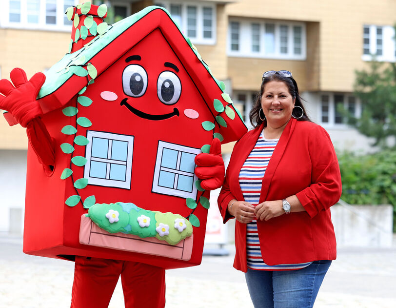 Vizebürgermeisterin und Wohnbaustadträtin Kathrin Gaál posiert neben dem Maskottchen für den Wiener Wohnbau, der Wohna Lisa. Das Kostüm der Wohna Lisa besteht aus einem roten Haus mit viel Blumenschmuck an den Fenstern, Augen, Mund, Armen und Beinen. 