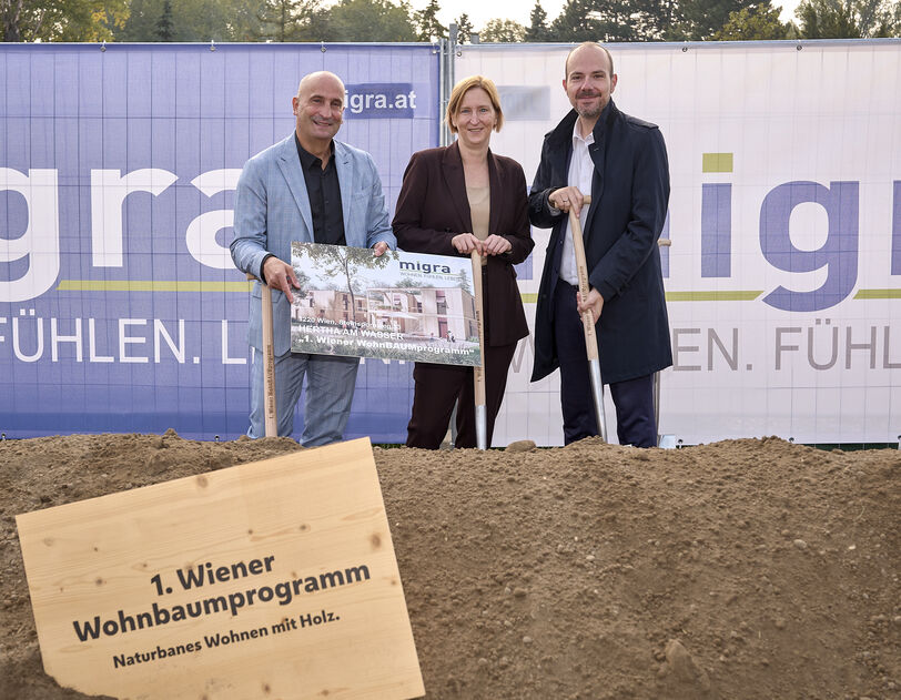 Drei Personen stehen auf einer Baustelle hinter einem Schild mit der Aufschrift '1. Wiener Wohnbaumprogramm Naturnahes Wohnen mit Holz' und halten ein Plakat vor einem Bauzaun mit dem Schriftzug 'migra.at'