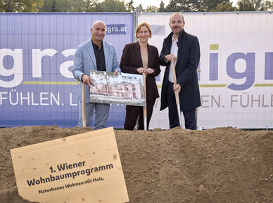 Drei Personen stehen auf einer Baustelle hinter einem Schild mit der Aufschrift '1. Wiener Wohnbaumprogramm Naturnahes Wohnen mit Holz' und halten ein Plakat vor einem Bauzaun mit dem Schriftzug 'migra.at'