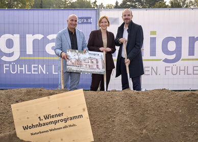 Drei Personen stehen auf einer Baustelle hinter einem Schild mit der Aufschrift '1. Wiener Wohnbaumprogramm Naturnahes Wohnen mit Holz' und halten ein Plakat vor einem Bauzaun mit dem Schriftzug 'migra.at'