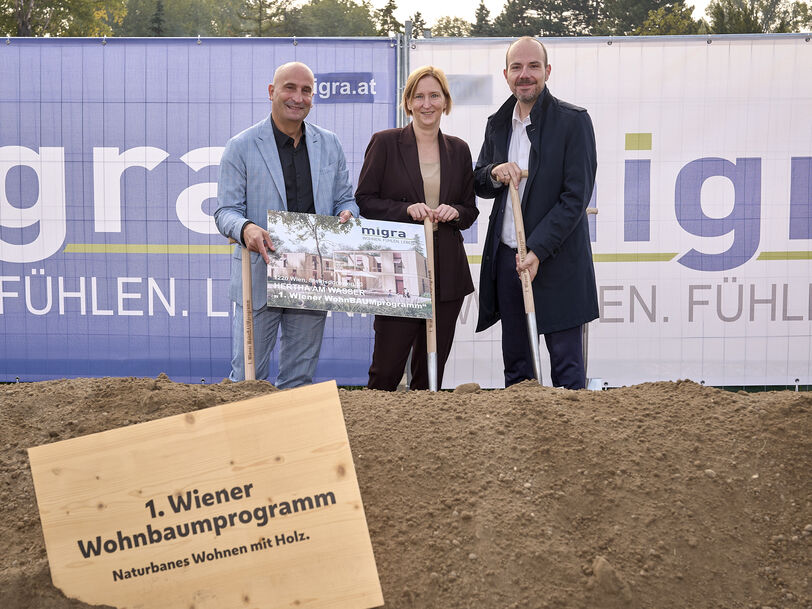 Drei Personen stehen auf einer Baustelle hinter einem Schild mit der Aufschrift '1. Wiener Wohnbaumprogramm Naturnahes Wohnen mit Holz' und halten ein Plakat vor einem Bauzaun mit dem Schriftzug 'migra.at'