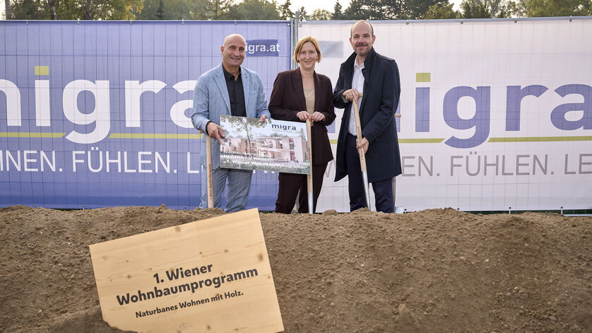 Drei Personen stehen auf einer Baustelle hinter einem Schild mit der Aufschrift '1. Wiener Wohnbaumprogramm Naturnahes Wohnen mit Holz' und halten ein Plakat vor einem Bauzaun mit dem Schriftzug 'migra.at'