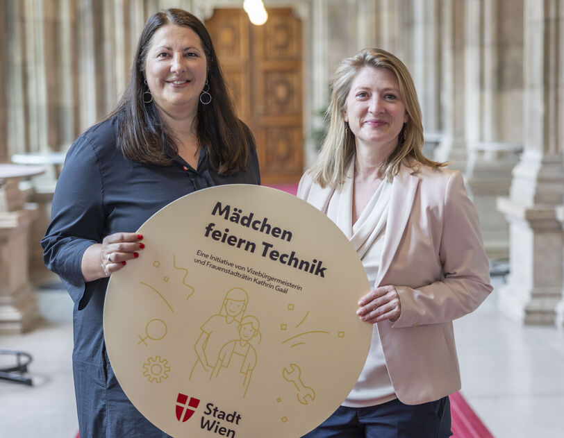 Vizebürgermeisterin und Frauenstadträtin Kathrin Gaál und Vizebürgermeisterin und Bildungsstadträtin Bettina Emmerling präsentieren die „Mädchen feiern Technik“-Workshops mit einem runden Schild, das von beiden gemeinsam gehaltne wird. Das Bild ist in den Arkadengängen des Rathauses aufgenommen worden. Kathrin Gaál trägt ein dunkelblaues Kleid, Bettina Emmerling eine dunkelblaue Hoseein beiges Oberteil mit Schalkragen und einen blassrosa Blazer darüber. Beide tragen ihre Haare offen.