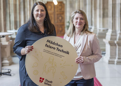 Vizebürgermeisterin und Frauenstadträtin Kathrin Gaál und Vizebürgermeisterin und Bildungsstadträtin Bettina Emmerling präsentieren die „Mädchen feiern Technik“-Workshops mit einem runden Schild, das von beiden gemeinsam gehaltne wird. Das Bild ist in den Arkadengängen des Rathauses aufgenommen worden. Kathrin Gaál trägt ein dunkelblaues Kleid, Bettina Emmerling eine dunkelblaue Hoseein beiges Oberteil mit Schalkragen und einen blassrosa Blazer darüber. Beide tragen ihre Haare offen.
