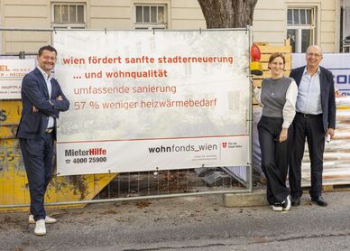 Drei Personen stehen vor einem Bauzaun mit einem Banner, das Informationen zur Stadtentwicklung und Wohnqualität in Wien zeigt