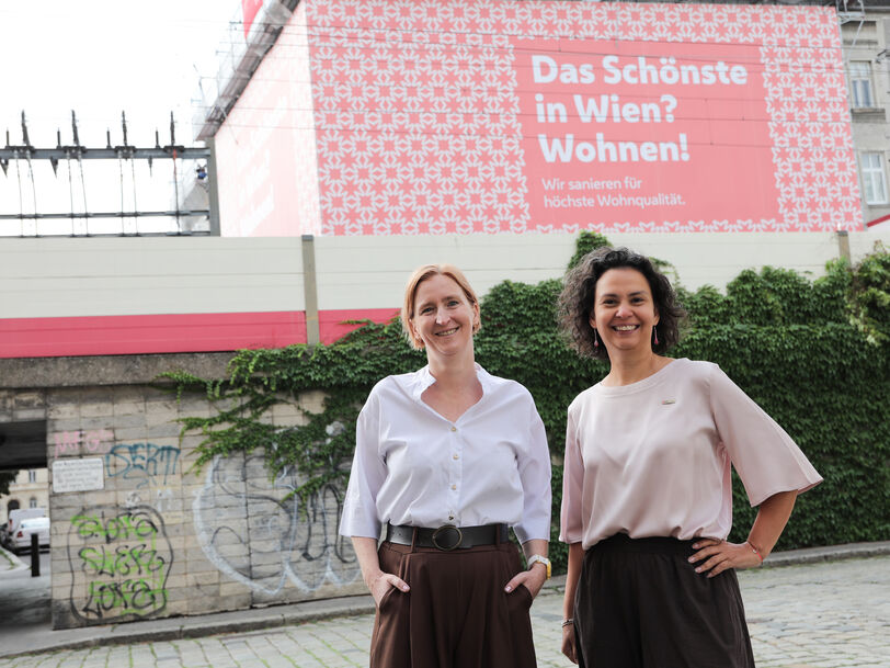 Zwei Frauen stehen vor einer mit Graffiti besprühten Mauer und einem Plakat mit der Aufschrift 'Das Schönste in Wien? Wohnen!'