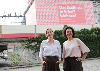 Zwei Frauen stehen vor einer mit Graffiti besprühten Mauer und einem Plakat mit der Aufschrift 'Das Schönste in Wien? Wohnen!'