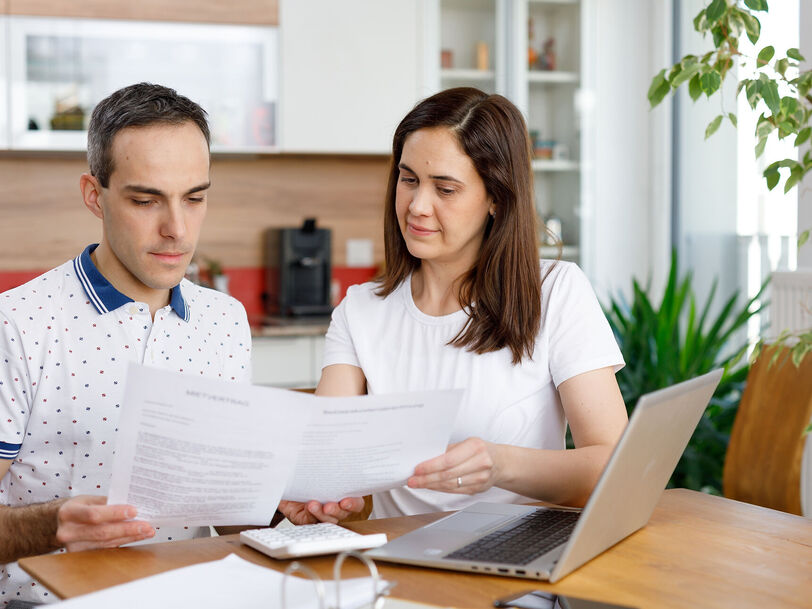 2 Personen sitzen an einem Holztisch und haben ein Notebook vor sich stehen. Zusätzlich hantieren sie mit einigen Zetteln und schauen diese konzentriert an. Im Hintergrund ist eine Küche erkennbar.