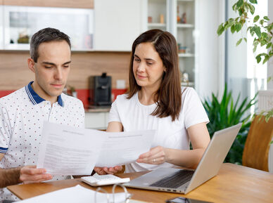 2 Personen sitzen an einem Holztisch und haben ein Notebook vor sich stehen. Zusätzlich hantieren sie mit einigen Zetteln und schauen diese konzentriert an. Im Hintergrund ist eine Küche erkennbar.