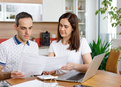 2 Personen sitzen an einem Holztisch und haben ein Notebook vor sich stehen. Zusätzlich hantieren sie mit einigen Zetteln und schauen diese konzentriert an. Im Hintergrund ist eine Küche erkennbar.