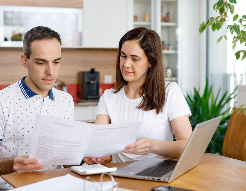 2 Personen sitzen an einem Holztisch und haben ein Notebook vor sich stehen. Zusätzlich hantieren sie mit einigen Zetteln und schauen diese konzentriert an. Im Hintergrund ist eine Küche erkennbar.