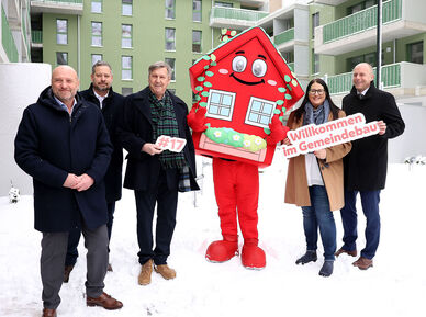 5 Personen stehen in winterlicher Business-Bekleidung im verschneiten Innenhof einer Neubauanlage. Ihnen zur Seite wurde wurde Wohna Lisa, das Maskottchen des geförderten Wohnbaus in Wien. Es ist eine als Haus verleidete rote Figur in Form eines Hauses mit Satteldach, großen Augen und einem freundlichen Lächeln.