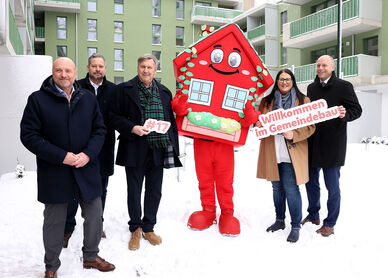 5 Personen stehen in winterlicher Business-Bekleidung im verschneiten Innenhof einer Neubauanlage. Ihnen zur Seite wurde wurde Wohna Lisa, das Maskottchen des geförderten Wohnbaus in Wien. Es ist eine als Haus verleidete rote Figur in Form eines Hauses mit Satteldach, großen Augen und einem freundlichen Lächeln.