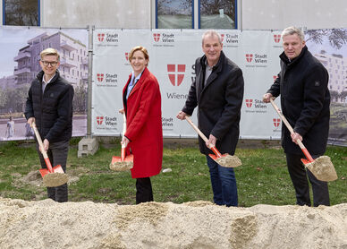4 Personen stehen mit Spaten mit Sand darauf hinter einem Sandhaufen und lächeln in die Kamera. I Hintergrund ist ein Bauzaun mit Transparenten auf denen Visualisierungen des Gemeindebau NEU und das Logo von Stadt Wien/Wiener Wohnen zu erkennen ist.