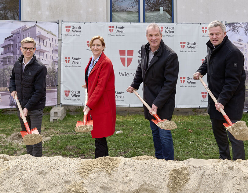 4 Personen stehen mit Spaten mit Sand darauf hinter einem Sandhaufen und lächeln in die Kamera. I Hintergrund ist ein Bauzaun mit Transparenten auf denen Visualisierungen des Gemeindebau NEU und das Logo von Stadt Wien/Wiener Wohnen zu erkennen ist.