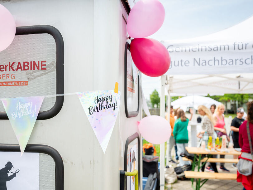Links im Vordergrund ist eine Telefonzelle zu erkennen, die zu einer Bücherkabine umgebaut wurde. Sie ist mit Luftballons und mit einer Wimpel-Girlande geschmückt, auf der Happy Birthday steht. Im Hintergrund sind Menschen bei und unter einem wohnpartner-Pavillionzelt zu erkennen. auf einem Heurigentisch stehen Saftboxen. 