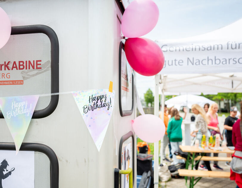Links im Vordergrund ist eine Telefonzelle zu erkennen, die zu einer Bücherkabine umgebaut wurde. Sie ist mit Luftballons und mit einer Wimpel-Girlande geschmückt, auf der Happy Birthday steht. Im Hintergrund sind Menschen bei und unter einem wohnpartner-Pavillionzelt zu erkennen. auf einem Heurigentisch stehen Saftboxen. 