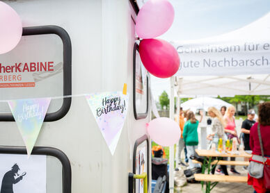 Links im Vordergrund ist eine Telefonzelle zu erkennen, die zu einer Bücherkabine umgebaut wurde. Sie ist mit Luftballons und mit einer Wimpel-Girlande geschmückt, auf der Happy Birthday steht. Im Hintergrund sind Menschen bei und unter einem wohnpartner-Pavillionzelt zu erkennen. auf einem Heurigentisch stehen Saftboxen. 