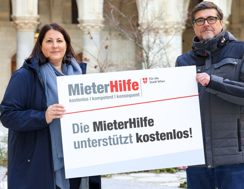 Kathrin Gaál und Christian Bartok stehen im Innenhof des Rathauses und halten gemeinsam eine Tafel auf der das Logo der MieterHilfe abgebildet ist und folgender Spruch zu lesen ist: "Die MieterHilfe unterstützt kostenlos!". Die beiden sind winterlich gekleidet.