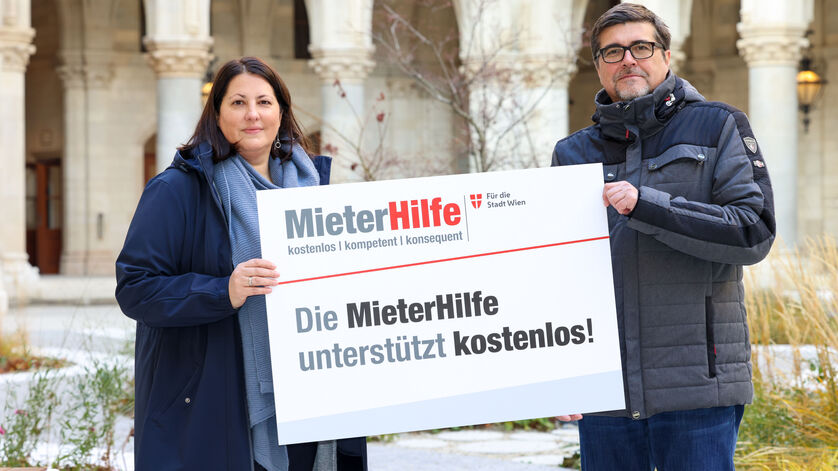 Kathrin Gaál und Christian Bartok stehen im Innenhof des Rathauses und halten gemeinsam eine Tafel auf der das Logo der MieterHilfe abgebildet ist und folgender Spruch zu lesen ist: "Die MieterHilfe unterstützt kostenlos!". Die beiden sind winterlich gekleidet.