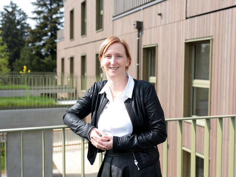Frau mit schwarzer Lederjacke und weißem Hemd lehnt an einem Geländer vor einem modernen Wohngebäude mit Holzfassade