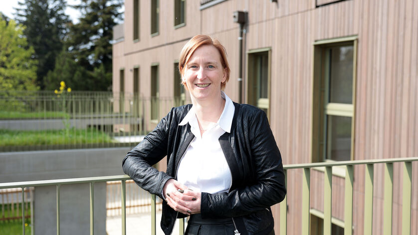 Frau mit schwarzer Lederjacke und weißem Hemd lehnt an einem Geländer vor einem modernen Wohngebäude mit Holzfassade