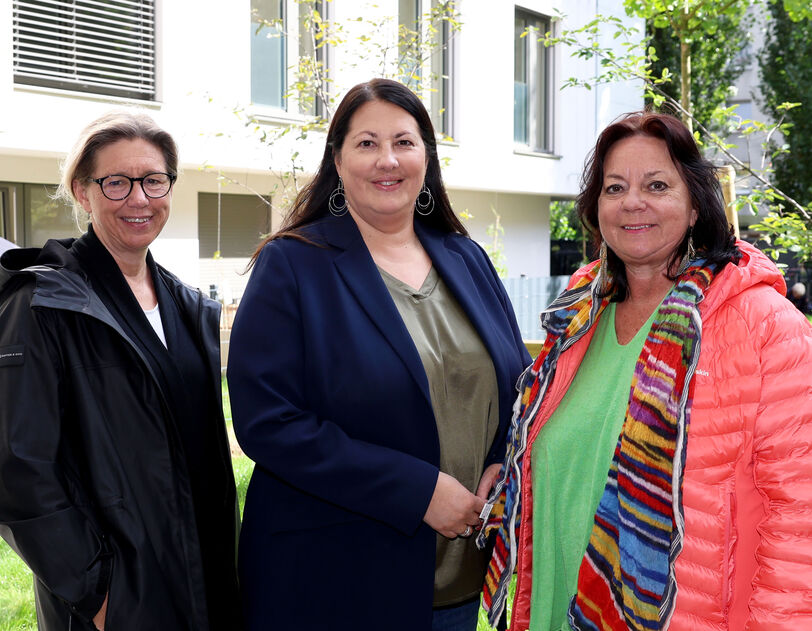 Wiener Wohnen Direktorin Karin Ramser, Vizebürgermeisterin und Wohnbaustadträtin Kathrin Gaál und wohnpartner-Bereichsleiterin Claudia Huemer stehen im begrünten Innenhof des neuen Gemeindebaus. Die drei sind trotz des offensichtlich freundlichen Wetters herbstlich gekleidet und lächeln freundlich in die Kamera.