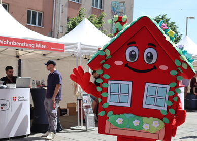 Das wienwohntbesser-Maskottchen Wohna Lisa, ein rotes Haus mit Armen und Beinen, einem Satteldach, einem freundlichen Lächeln im Gesicht und mit ausreichend Blumenschmuck steht auf einem Platz und winkt in die Kamera. Im Hintergrund ist ein Pagodenzelt sichtbar in dem an einem Pult mit Laptops darauf, zwei Personen eine weitere Person beraten, die davor steht.