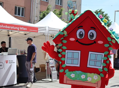 Das wienwohntbesser-Maskottchen Wohna Lisa, ein rotes Haus mit Armen und Beinen, einem Satteldach, einem freundlichen Lächeln im Gesicht und mit ausreichend Blumenschmuck steht auf einem Platz und winkt in die Kamera. Im Hintergrund ist ein Pagodenzelt sichtbar in dem an einem Pult mit Laptops darauf, zwei Personen eine weitere Person beraten, die davor steht.