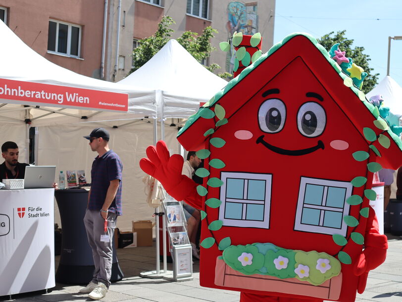 Das wienwohntbesser-Maskottchen Wohna Lisa, ein rotes Haus mit Armen und Beinen, einem Satteldach, einem freundlichen Lächeln im Gesicht und mit ausreichend Blumenschmuck steht auf einem Platz und winkt in die Kamera. Im Hintergrund ist ein Pagodenzelt sichtbar in dem an einem Pult mit Laptops darauf, zwei Personen eine weitere Person beraten, die davor steht.