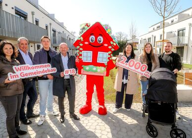 Sieben Personen stehen im Innenhof einer Wohnhausanlage. Die Anlage ist neu und hat drei Geschoße. In der Mitte der Gruppe steht das Maskottchen des Wiener Wohnbaus, Wohna Lisa. Sie ist ein rotes freundlich dreinblickendes Haus mit Satteldach auf zwei Beinen. Die Personen halten die Schriftzüge "Willkommen im Gemeindebau", "#18" und "2.000ste Gemeindewohnung NEU" in der Hand. Vor zwei Personen steht ein Kinderwagen. 