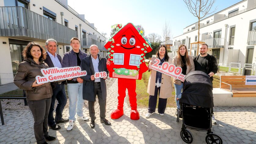 Sieben Personen stehen im Innenhof einer Wohnhausanlage. Die Anlage ist neu und hat drei Geschoße. In der Mitte der Gruppe steht das Maskottchen des Wiener Wohnbaus, Wohna Lisa. Sie ist ein rotes freundlich dreinblickendes Haus mit Satteldach auf zwei Beinen. Die Personen halten die Schriftzüge "Willkommen im Gemeindebau", "#18" und "2.000ste Gemeindewohnung NEU" in der Hand. Vor zwei Personen steht ein Kinderwagen. 