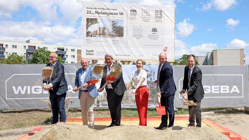 Sechs Personen mit Spaten stehen auf einer Baustelle vor einem großen Schild und schaufeln Erde in eine Sandkiste