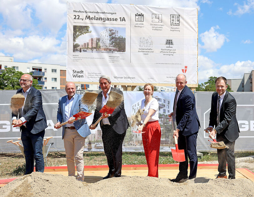 Sechs Personen mit Spaten stehen auf einer Baustelle vor einem großen Schild und schaufeln Erde in eine Sandkiste