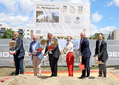 Sechs Personen mit Spaten stehen auf einer Baustelle vor einem großen Schild und schaufeln Erde in eine Sandkiste