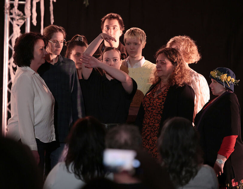 Mehrere Personen stehen knapp beisammen und blicken auf eine in der Mitte platzierte Person die mit ihren Armen die Äste eines Baumes darstellt. Der Hintergrund ist schwarz, im Vordergrund erkennt man Personen sitzend von hinten, die auf die Personengruppe schauen. 