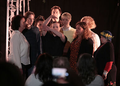 Mehrere Personen stehen knapp beisammen und blicken auf eine in der Mitte platzierte Person die mit ihren Armen die Äste eines Baumes darstellt. Der Hintergrund ist schwarz, im Vordergrund erkennt man Personen sitzend von hinten, die auf die Personengruppe schauen. 