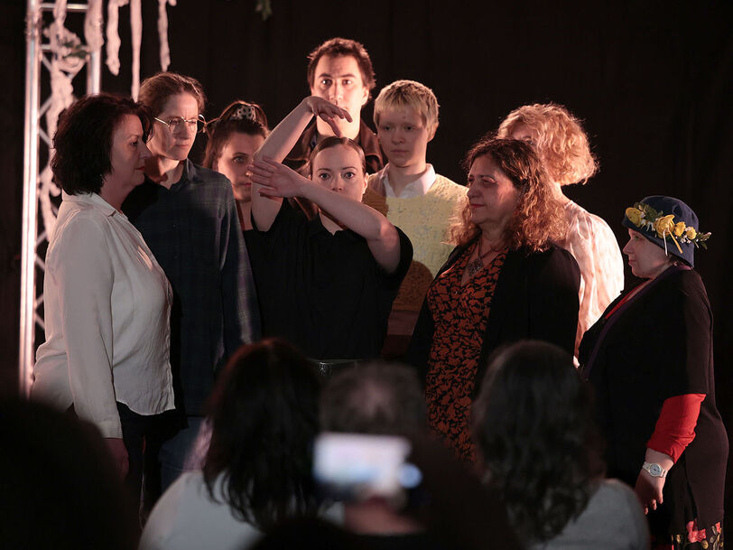 Mehrere Personen stehen knapp beisammen und blicken auf eine in der Mitte platzierte Person die mit ihren Armen die Äste eines Baumes darstellt. Der Hintergrund ist schwarz, im Vordergrund erkennt man Personen sitzend von hinten, die auf die Personengruppe schauen. 