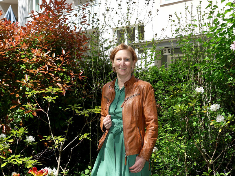 Frau in grünem Kleid und brauner Lederjacke steht in einem üppigen Garten vor einem mehrstöckigen Wohnhaus