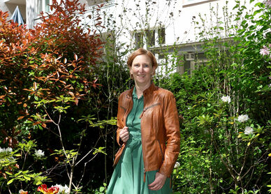 Frau in grünem Kleid und brauner Lederjacke steht in einem üppigen Garten vor einem mehrstöckigen Wohnhaus