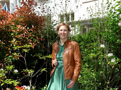 Frau in grünem Kleid und brauner Lederjacke steht in einem üppigen Garten vor einem mehrstöckigen Wohnhaus