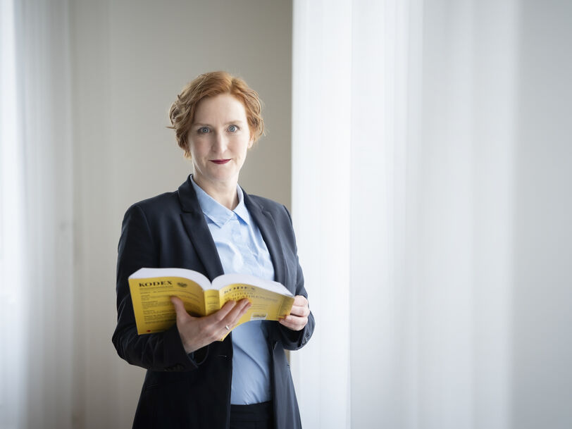Frau mit roten Haaren in dunklem Blazer und hellblauer Bluse steht vor hellem Vorhang und hält ein aufgeschlagenes gelbes Buch, den Mietrechts-Kodex, in den Händen