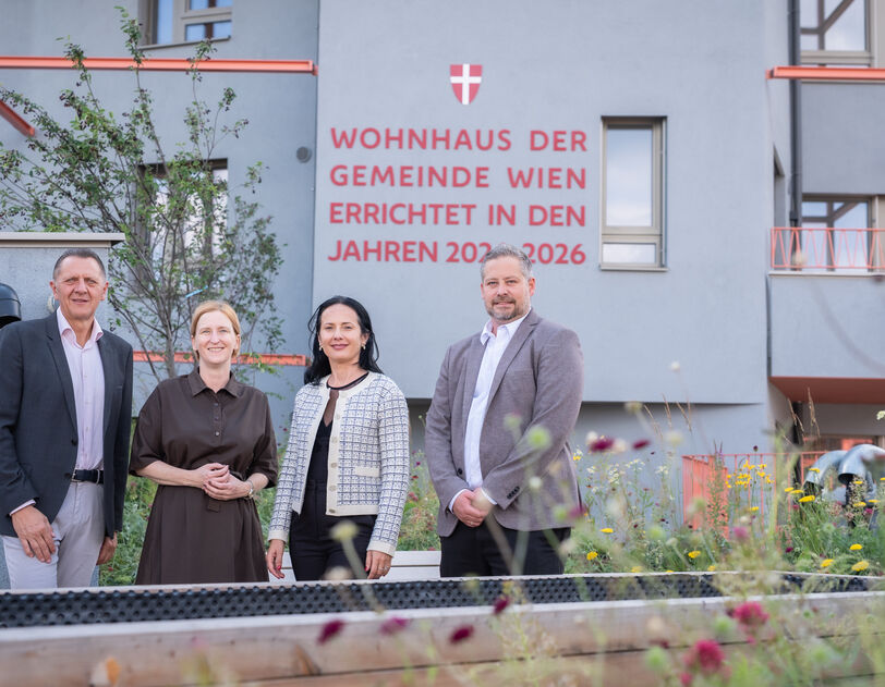 Vier Personen stehen vor einem modernen Wohnhaus mit der Aufschrift 'Wohnhaus der Gemeinde Wien errichtet in den Jahren 2023-2026'