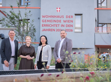 Vier Personen stehen vor einem modernen Wohnhaus mit der Aufschrift 'Wohnhaus der Gemeinde Wien errichtet in den Jahren 2023-2026'