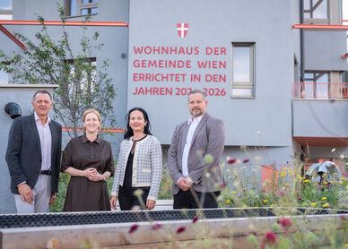 Vier Personen stehen vor einem modernen Wohnhaus mit der Aufschrift 'Wohnhaus der Gemeinde Wien errichtet in den Jahren 2023-2026'