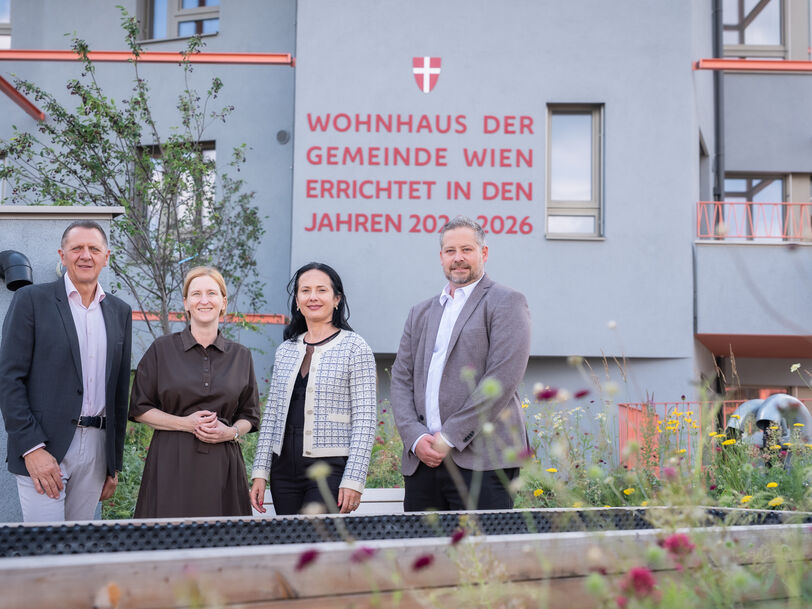 Vier Personen stehen vor einem modernen Wohnhaus mit der Aufschrift 'Wohnhaus der Gemeinde Wien errichtet in den Jahren 2023-2026'
